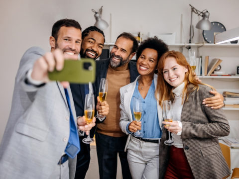 Group of 5 people holding champagne flutes and taking a picture in an office