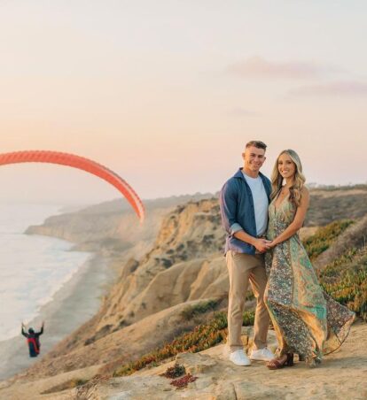 a couple standing on a cliff above the ocean with a parasailer in the background