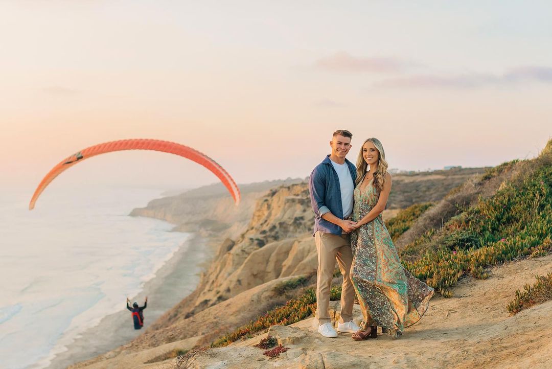 a couple standing on a cliff above the ocean with a parasailer in the background