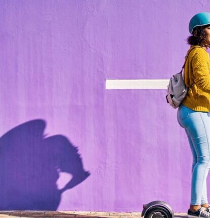 woman in a yellow sweater riding a scooter past a bright purple wall