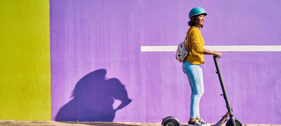 woman in a yellow sweater riding a scooter past a bright purple wall
